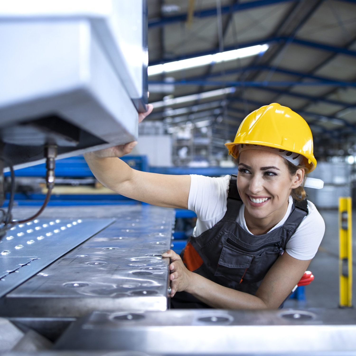 Female factory worker in protective uniform and hardhat operating industrial machine at production line.